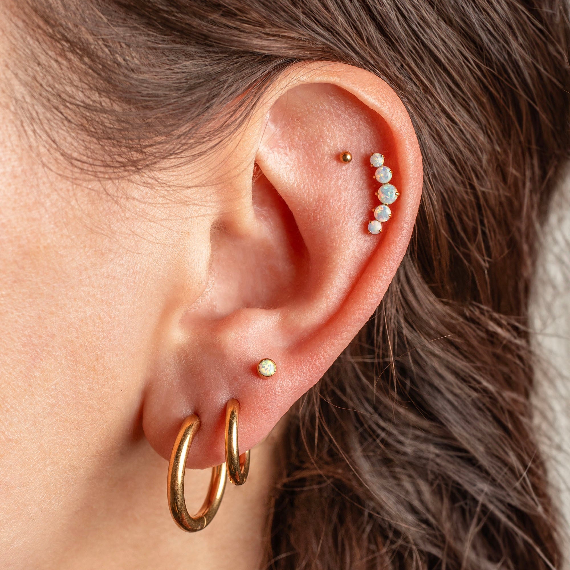 Close-up of a person's ear showcasing multiple gold and pearl ear piercings, including hoop earrings, a stud, and a curved row of small white pearls on the helix.