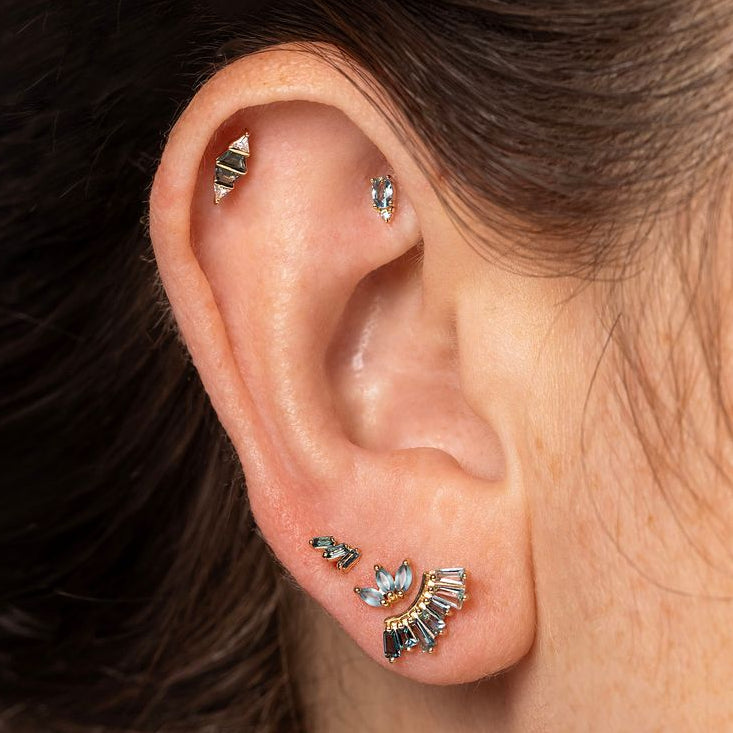 Close-up of a person's ear showcasing multiple small silver-toned ear piercings, including a helix stud, lobe studs, and a cluster of three small wing-shaped studs on the cartilage.