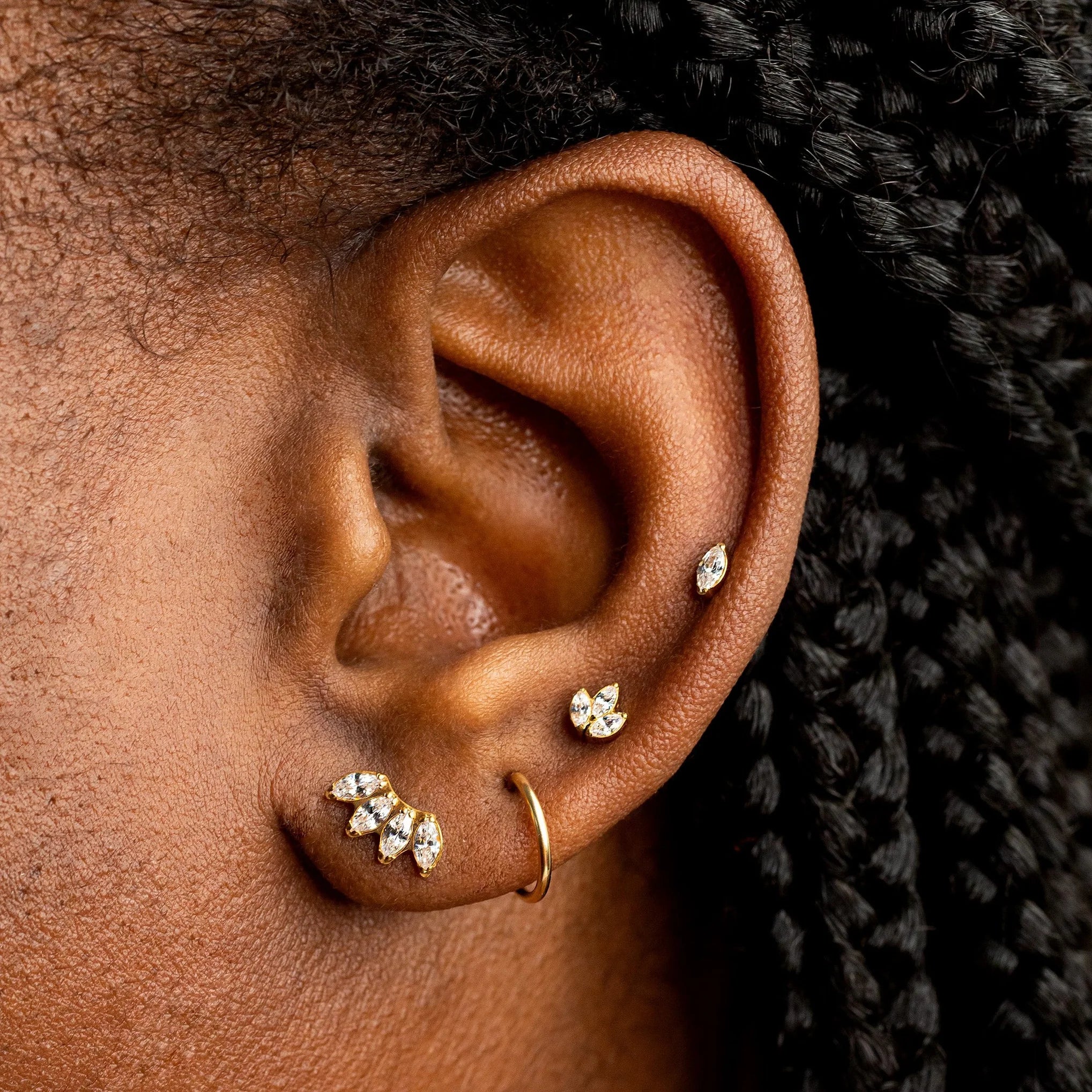 Close-up of an ear wearing multiple gold ear piercings, including a hoop in the cartilage, a small stud, a cluster stud, and a curved barbell, with dark braided hair visible.