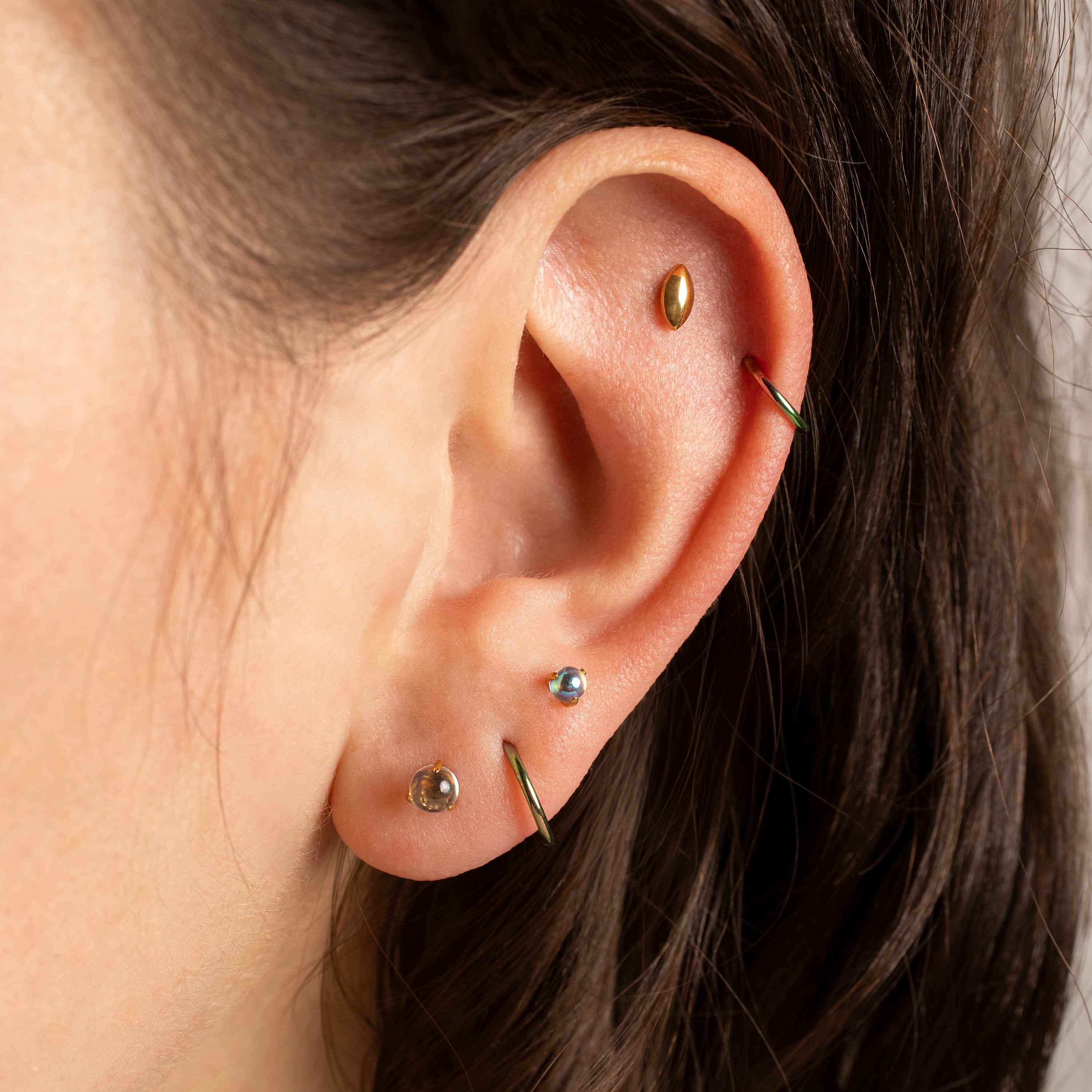 Close-up of a person's ear showcasing multiple piercings, including a gold hoop in the lobe, a gold helix, a silver cartilage stud, and a green curved barbell.