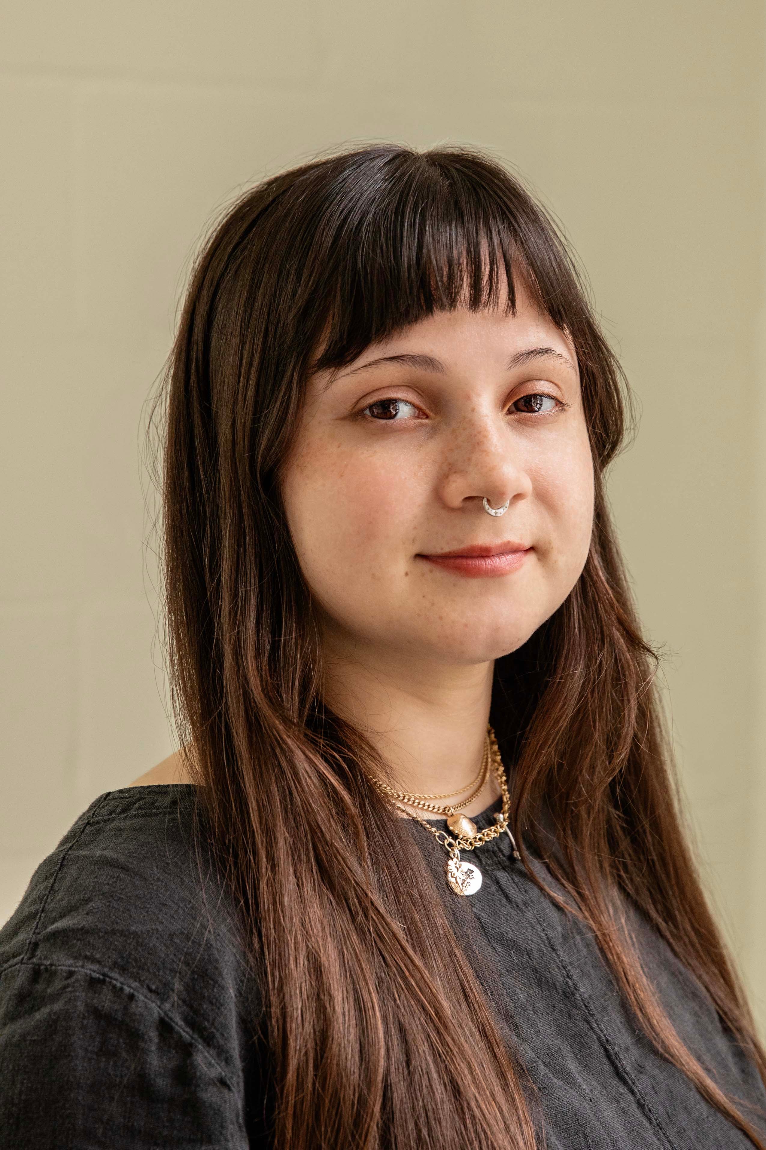 Woman with long brown hair and a nose ring, wearing a dark shirt against a beige background