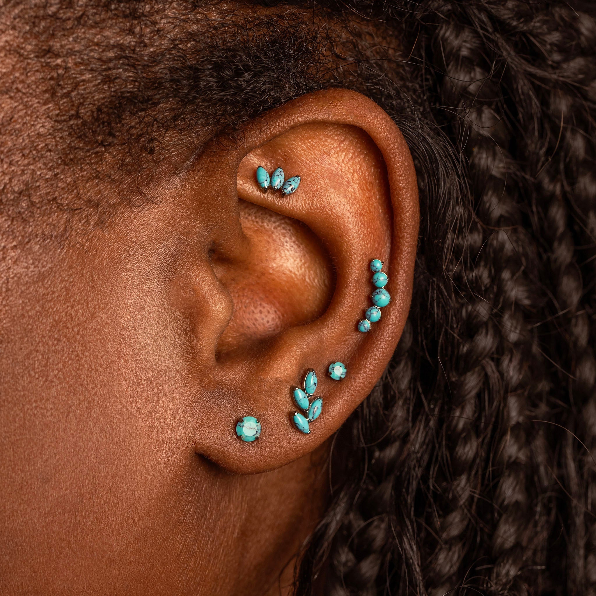 Close-up of a person's ear adorned with multiple turquoise-colored stud and cartilage earrings, with dark braided hair visible.